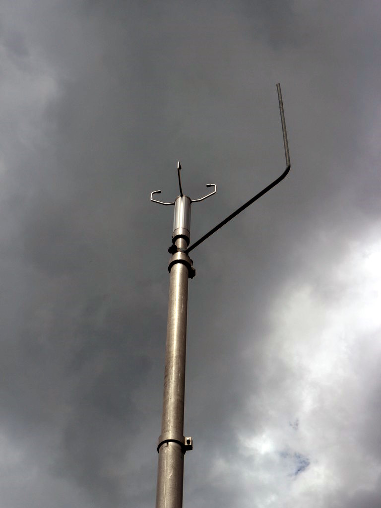 Die Abbildung zeigt ein Wind-Ultraschall-Anemometer, welches auf einem 10 m hohen Mast angebracht ist. Neben dem Anemometer ist ein Blitzableiter installiert.