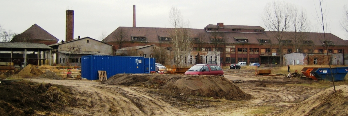 Im Hintergrund befinden sich alte zerfallene Industriegebäude in Bernau. Im Vordergrund steht ein blauer Container. Der Boden ist braun und zerfahren.