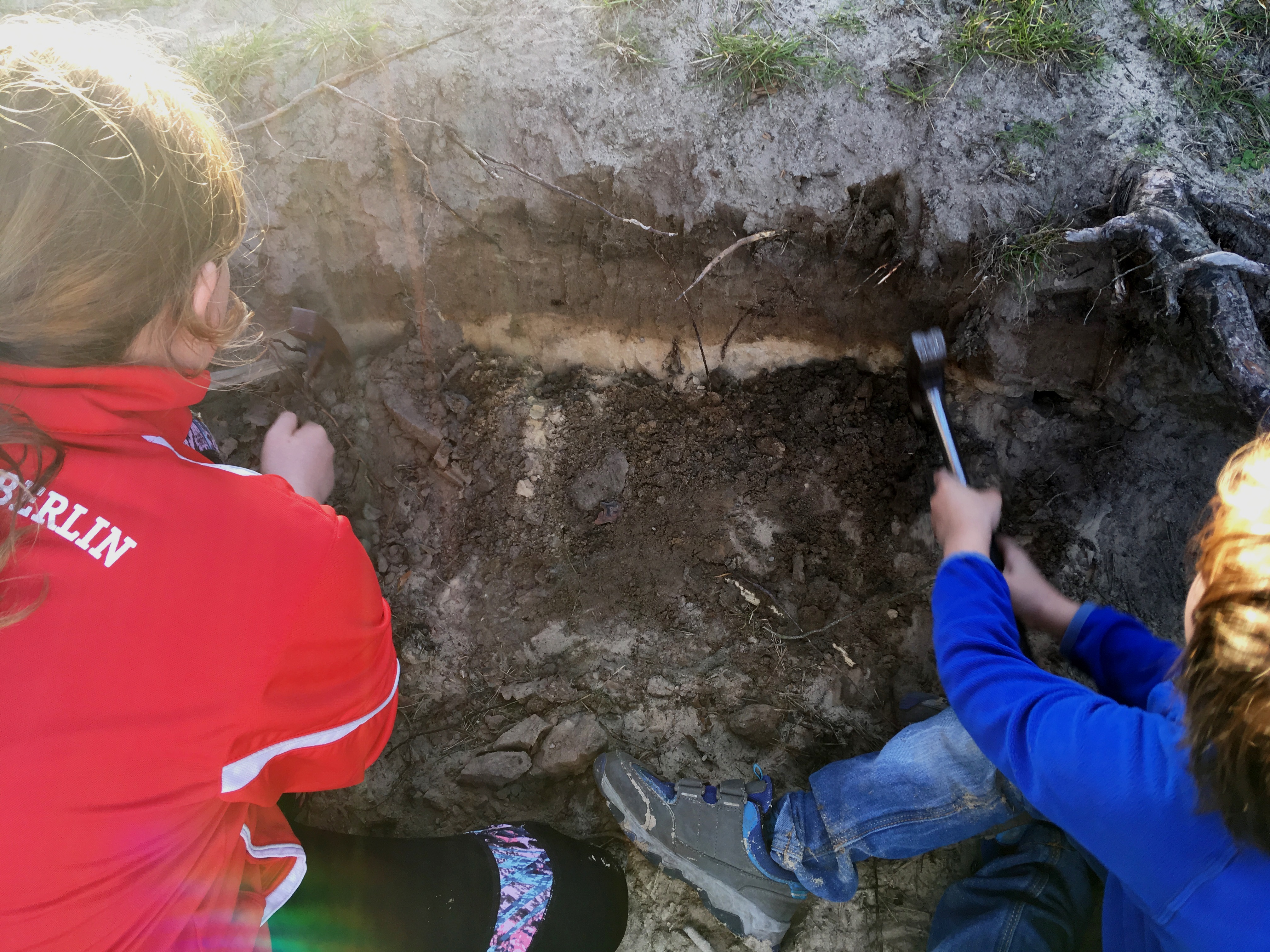 Zwei Kinder sitzen in einer Bodengrube und klopfen verschiedene Bodenschichten frei.