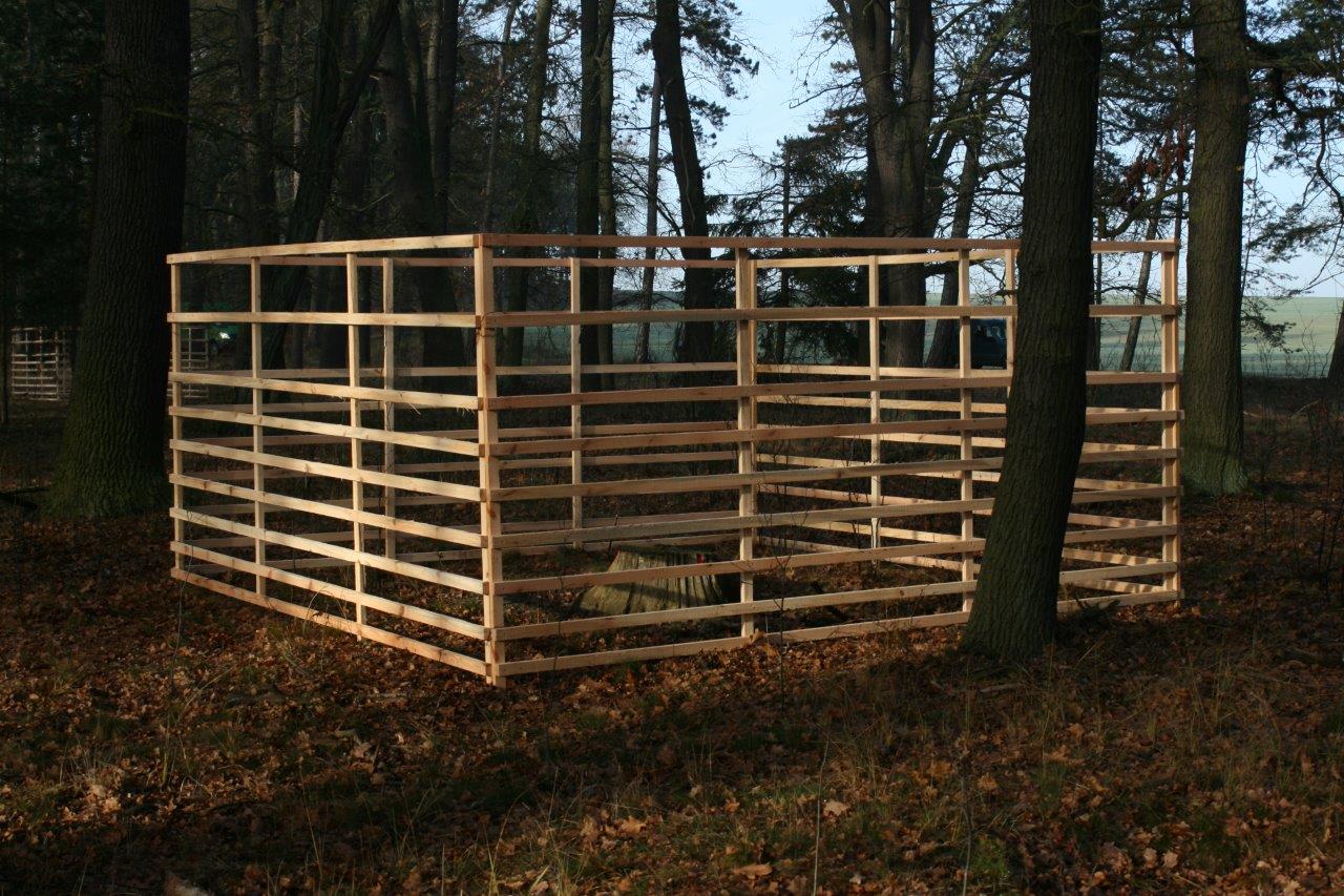 Ein aus Holz gebautes Hordengatter. Das Hordengatter steht im Wald zwischen mehreren Bäumen.