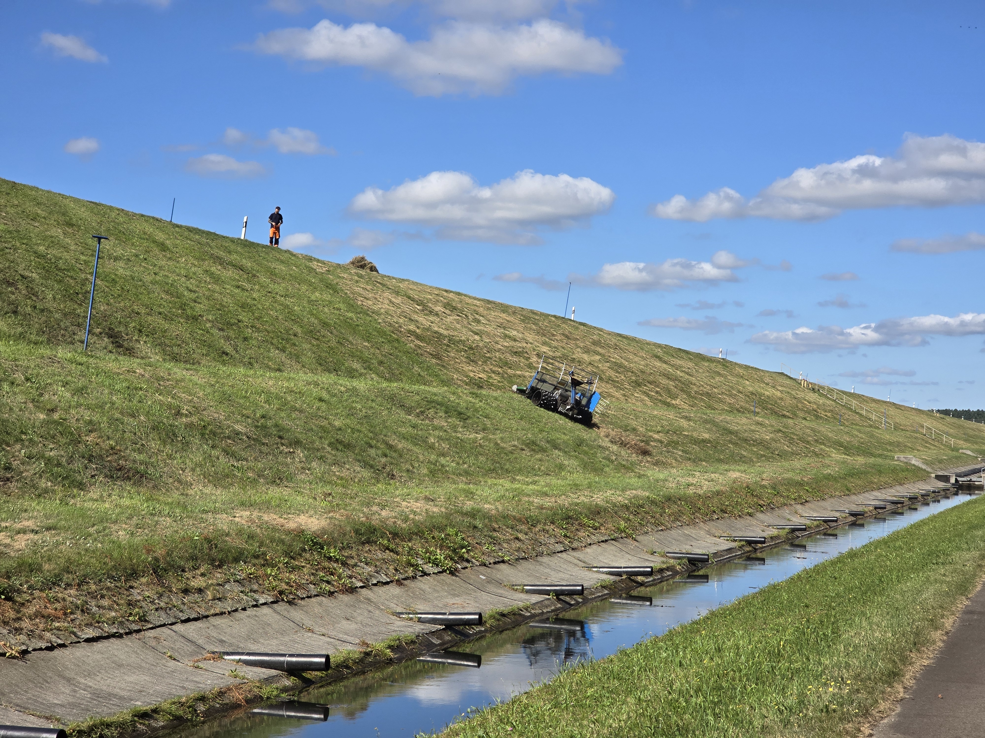 Mahd des Staudamms Damm der Talsperre nach rechts abfallend mit einem Betonentwässerungskanal davor. Der Hang ist mit saftig grünem Gras bewachsen, welches gerade gemäht wird. Auf dem Hang steht ein Arbeiter. Im Hintergrund strahlt der blaue Himmel mit nur wenigen vereinzelten Wolken.
