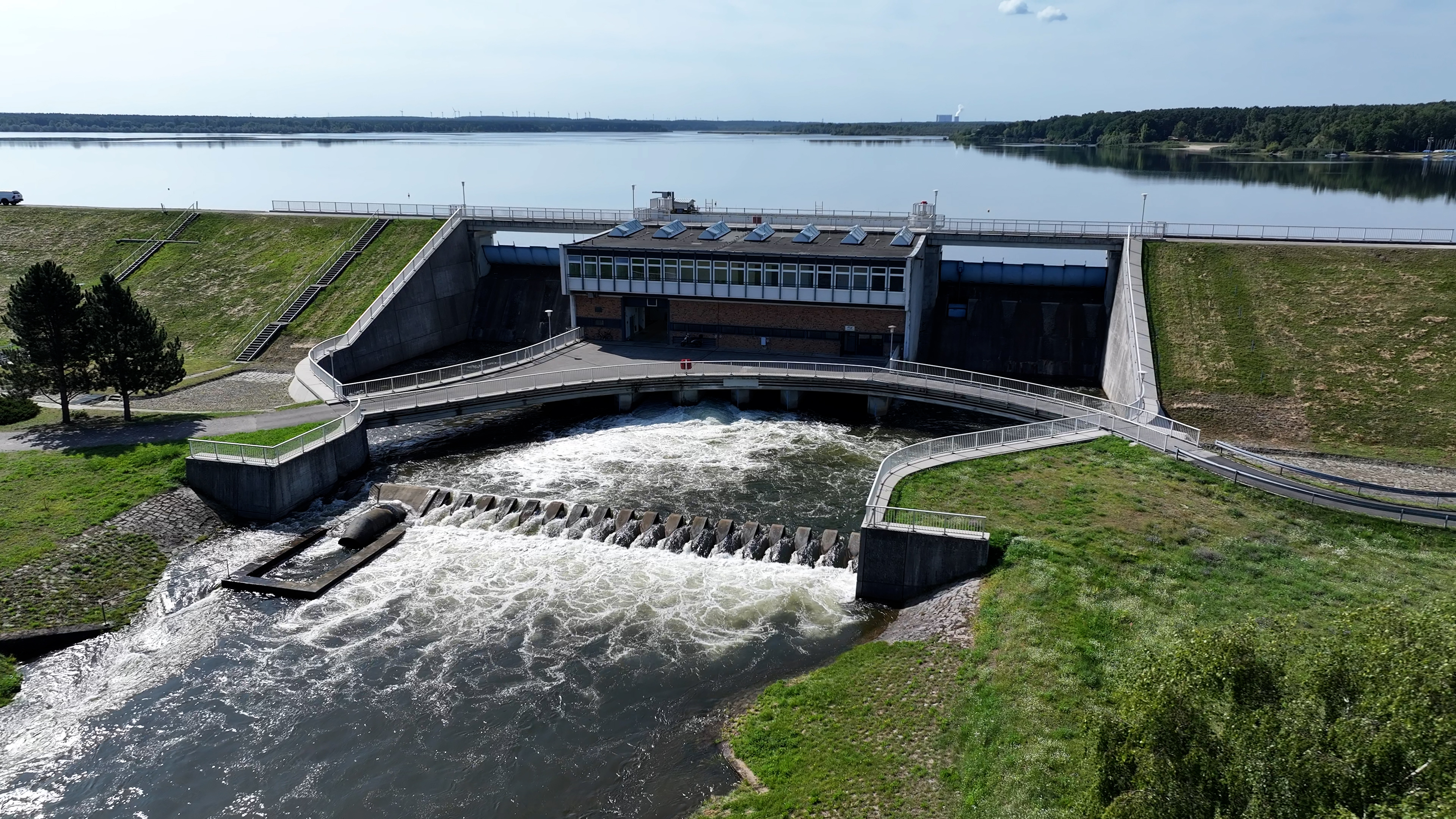 Das Grundablass- und Entlastungsbauwerk mit dem Tosbecken, im Hintergrund der Stausee Grundablass- und Entlastungsbauwerk der Talsperre Spremberg mit dem Tosbecken im Vordergrund, gesäumt von grünen Wiesen und links zwei Bäumen. Im Hintergrund ist das Wasser der Talsperre zu sehen, in welchem sich der blaue Himmel spiegelt.