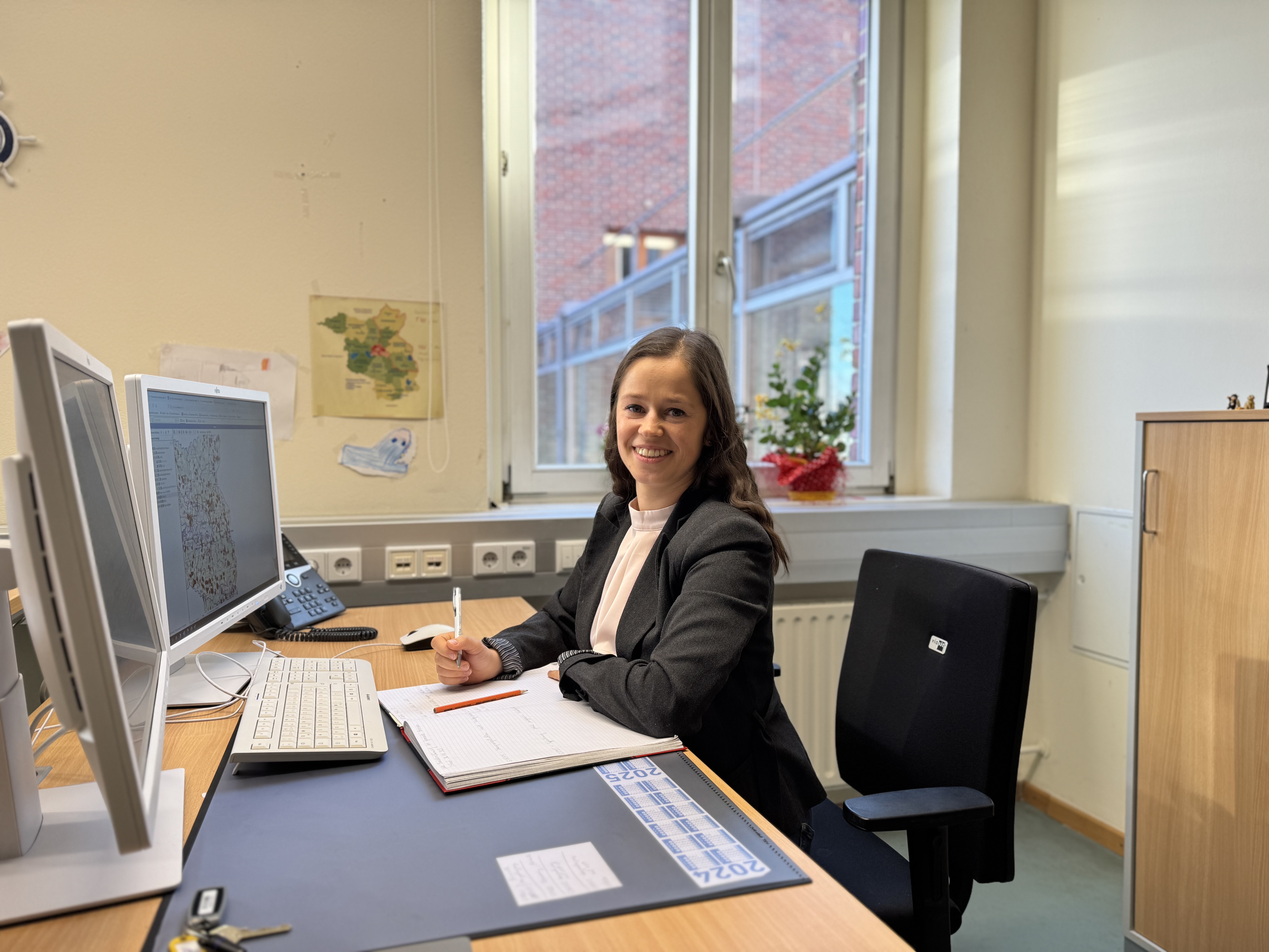 Frau Dobrzycka sitzt an einem Schreibtisch vor zwei Bildschirmen und lächelt in die Kamera. Im Hintergrund an der Wand sind Kindermalereien und eine Landkarte vom Land Brandenburg zu sehen. 
