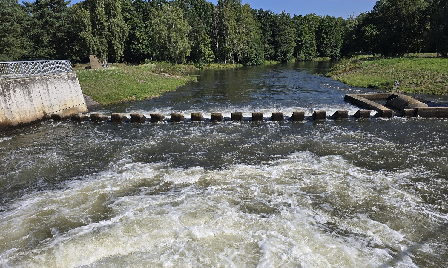 Übergang vom Tosbecken in die Spree Übergang vom Tosbecken in die Spree an der Talsperre Spremberg. Im Vordergrund ist das schäumende Wasser des Tosbeckens zu sehen, das durch die Struktur der Anlage aufgewühlt wird. Eine Reihe von Betonblöcken dient als Barriere, über die das Wasser fließt. Im Hintergrund fließt die Spree ruhig weiter. Uferböschungen mit üppigem Grün säumen den Flusslauf, und dahinter erstreckt sich ein dichter Wald, der die Szenerie einrahmt.