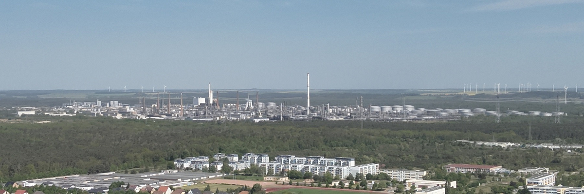Blick über eine Stadtlandschaft mit einem großen Industriegebiet und Windkraftanlagen am Horizont. Links im Vordergrund sind einige Häuser zu sehen.