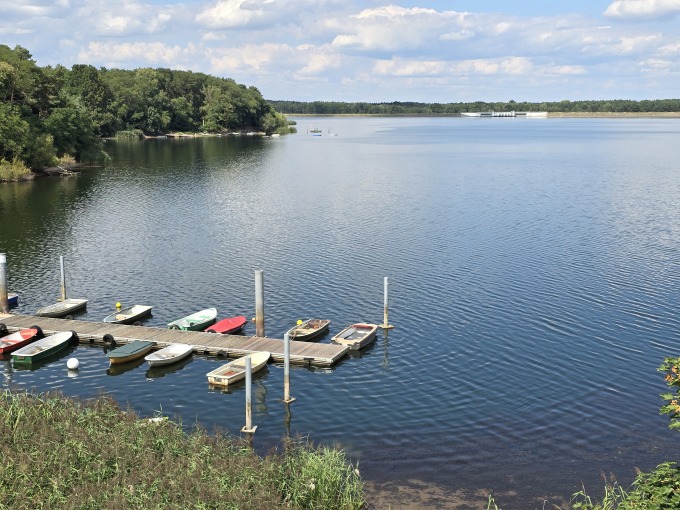 Landschaftsszene der Talsperre  im Bereich Spreecamp Groß Döbbern. Der See ist von beiden Seiten von üppigem Grün umgeben und im Wasser spiegelt sich der blaue Himmel mit vereinzelten Wolken. Im Vordergrund gibt es mehrere kleine Boote in verschiedenen Farben, die an einem Holzsteg festgemacht sind. Die Vegetation im Vordergrund besteht hauptsächlich aus Schilf.
