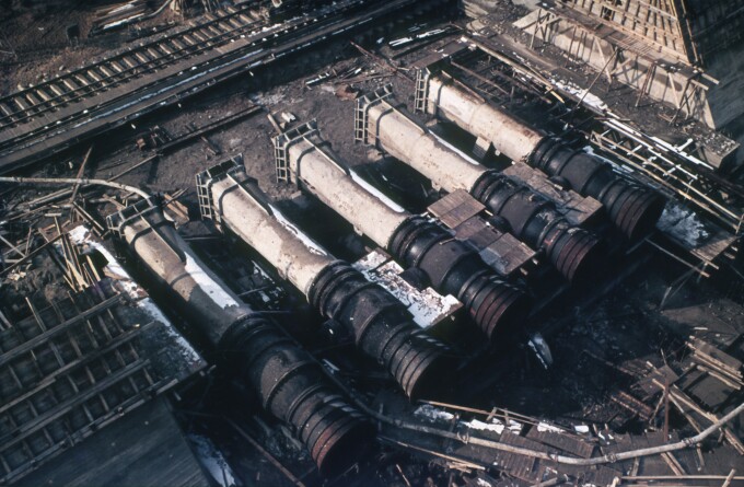Das Bild zeigt in grau-, brau- und schwarztönen, die im Bau befindlichen Grundablässe des Auslaufbauwerkes der Talsperre Spremberg aus der Vogelperspektive. 5 massive Rohre liegen parallel zueinander und sind Teil der komplexen Infrastruktur. Die Konstruktion wirkt unvollendet, umgeben von Gerüsten und Baumaterialien.