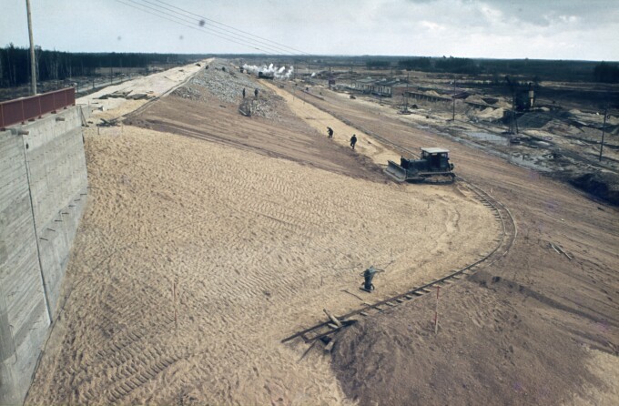 Sand- und Kieslandschaft aus der Zeit des Baus der Talsperre, links gesäumt mit einem Teil des neu gebauten Auslaufbauwerkes. Eine Planierraupe schiebt den Sand den Damm hinauf und einige Arbeiter sind im Hintergrund zu sehen.