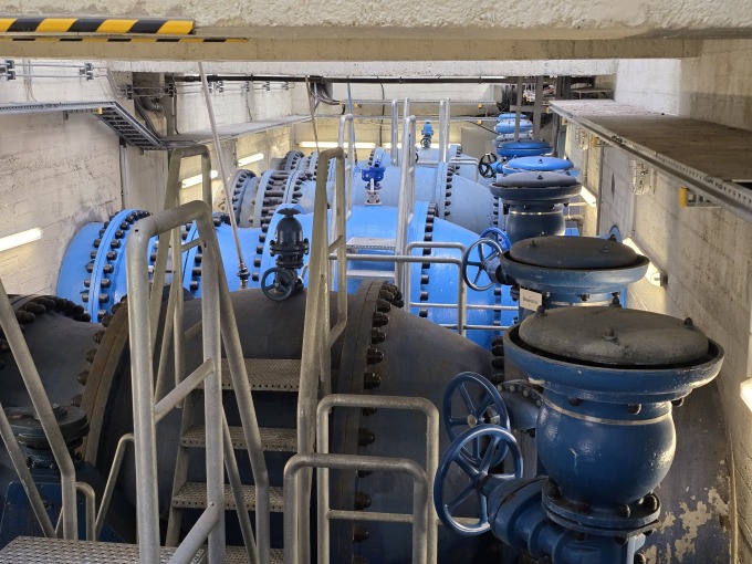 Heutige Innenansicht des Technikgebäudes der Talsperre Spremberg, speziell die fünf Grundablässe. Man sieht große, blaue Rohre und Ventile, die Teil des Steuerungssystems für den Wasserdurchfluss sind. Metalltreppen und -geländer ermöglichen den Zugang zur Wartung. Die betonierte Umgebung ist funktional und zeugt von der industriellen Notwendigkeit der Anlage. Die Beleuchtung betont die technischen Details und die Ordnung des Raumes. Insgesamt vermittelt das Bild einen Eindruck von der Komplexität und dem technischen Aufwand, der hinter der Wasserregulierung steckt.