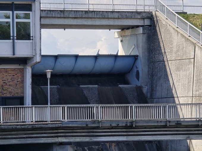 Das Bild zeigt einen Hochwasserüberfallrücken mit Fischbauchklappe an der Talsperre Spremberg. Die Aufnahme fängt die architektonische Stärke und Funktionalität dieser Wasserbauanlage ein. Der Fokus liegt auf der komplexen Struktur der Klappe und dem Betonbau, der die enormen Wassermassen kontrolliert. Die grauen Betonelemente dominieren das Bild und vermitteln ein Gefühl von Solidität und Dauerhaftigkeit. Das ablaufende Wasser bildet einen dynamischen Kontrast zu den statischen Elementen und unterstreicht die Kraft des Wassers.