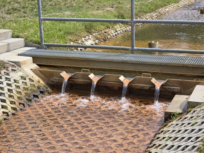 Das Bild zeigt quer durch das Bild verlaufend einen kleinen Entwässerungsgraben, der bräunliches Wasser führt. Geteilt wird der Graben durch das Messwehr, welches durch Betontreppen von beiden Böschungen aus begehbar ist. Über das Wehr führt ein Bediensteg mit Geländer. Im Hintergrund ist Wald zu sehen, welcher mit einem dunkelgrünen Zaun abgetrennt ist. 