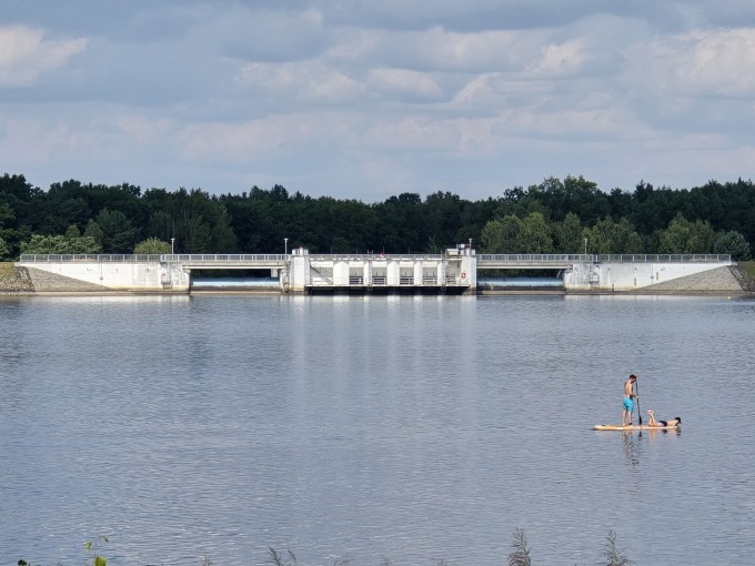 Das Bild zeigt in der unteren Hälfte ruhiges hellblaues Gewässer, in dem zwei Personen mit einem Stand-up-Paddle unterwegs sind. Das zentrale Merkmal dieses Bildes ist das Große Auslaufbauwerk in der Bildmitte, welches sich durch seine helle auszeichnet. Hier fließt das Wasser in den Spreelauf zurück. Im Hintergrund befindet sich Wald und am blauen Himmel einige Wolken.