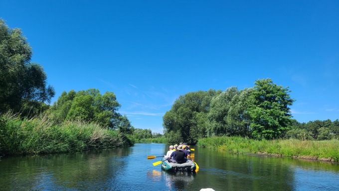 Ein Fluss, der links und rechts von mit Bäumen und Gebüsch bewachsenen Ufern eingerahmt wird, mittig ein Schlauchboot von hinten, in dem mehrere Personen sitzen und paddeln. 
