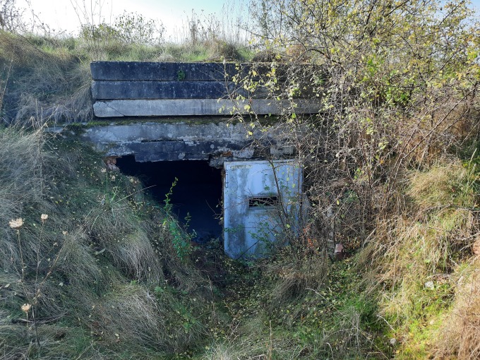 In einer kleinen Landschaftsmulde eingebaut, steht ein kleines Gebäude, dessen Tür offensteht und das durch Gräser und Gebüsch stark zugewachsen ist. 