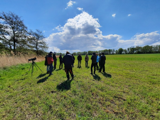 Eine im Kreis stehende Menschengruppe, die auf einer abgemähten grünen Wiesenfläche steht, im Hintergrund Baumreihen.